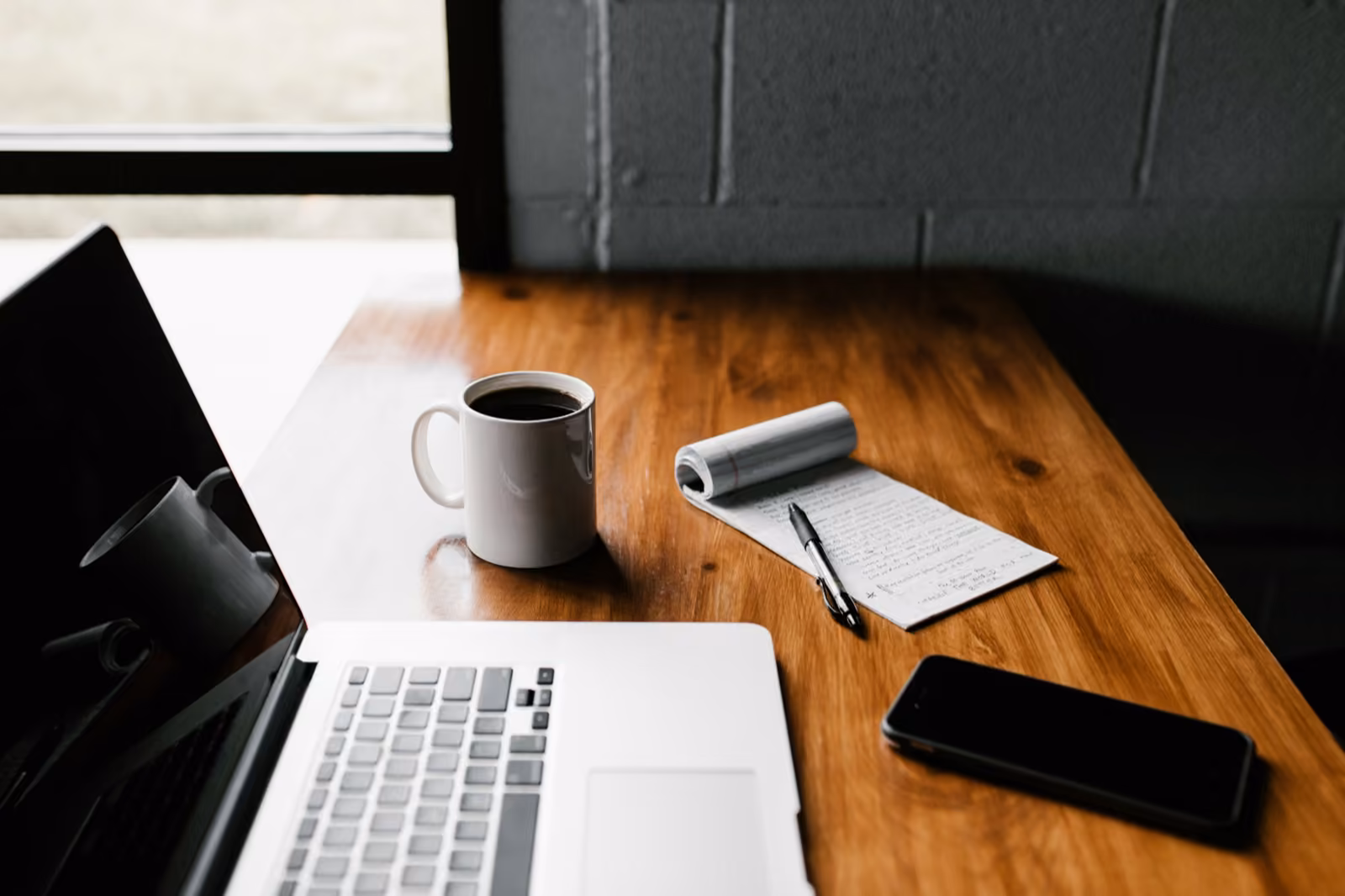 A MacBook, coffee, and a paper notebook on a wood table — the founder doing math.