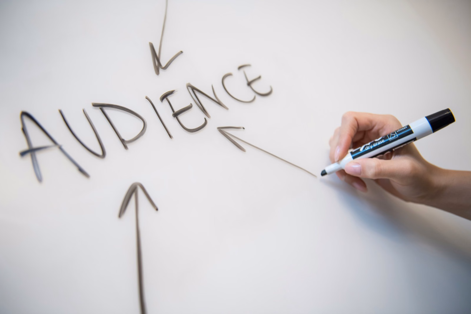 Hand writing the word Audience on a whiteboard with arrows converging on it, illustrating audience targeting strategy.